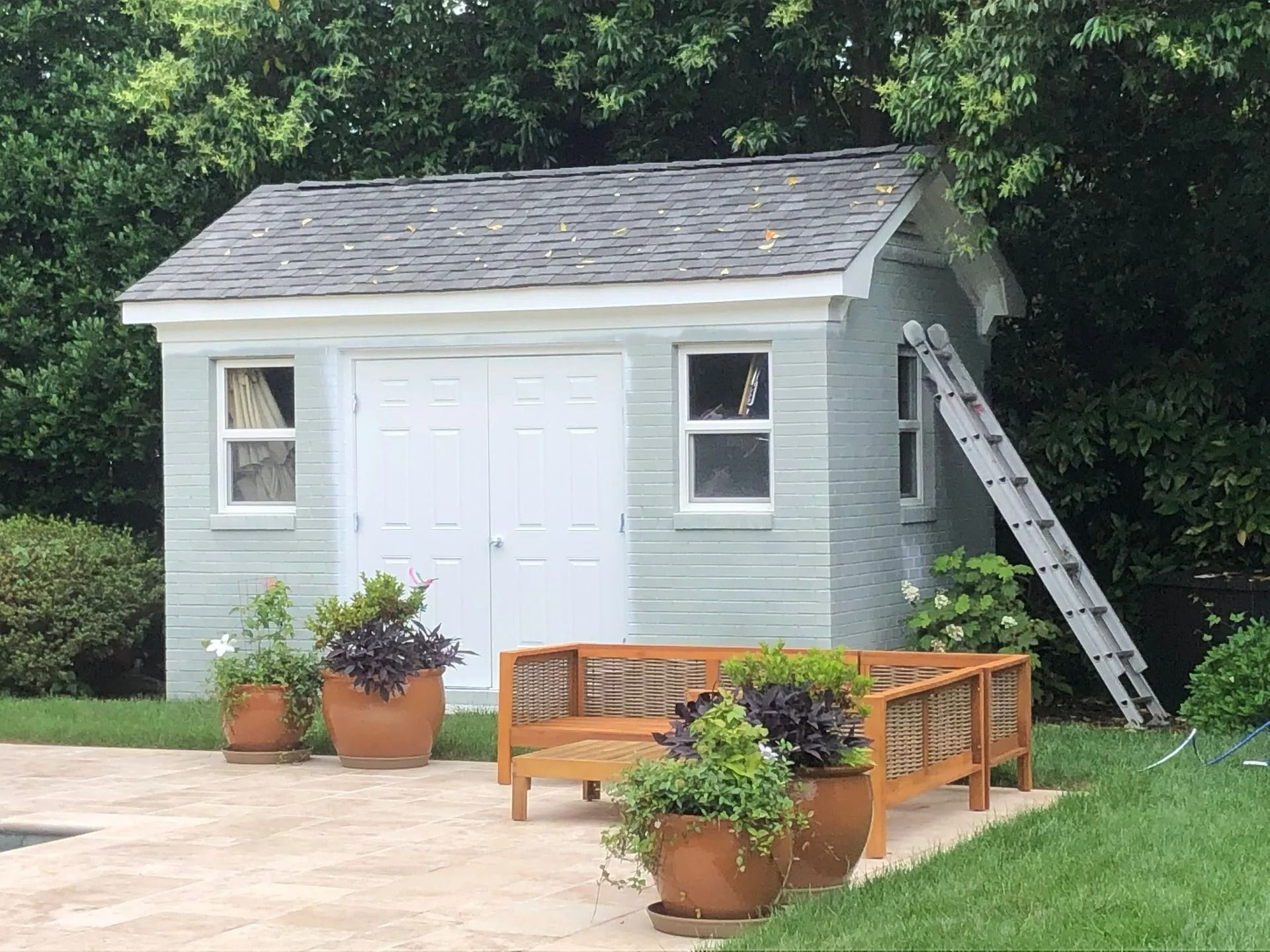 Shed with gray roof, white doors, and windows, next to a ladder. Includes bench, potted plants, on a stone patio.