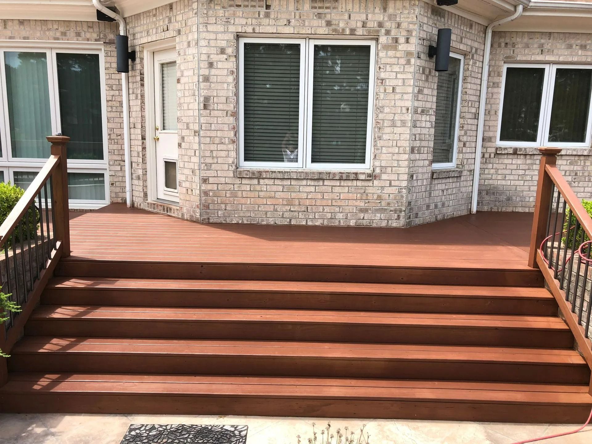Wooden deck with steps leading up to a brick house. Brown wood, white windows, and black railings.