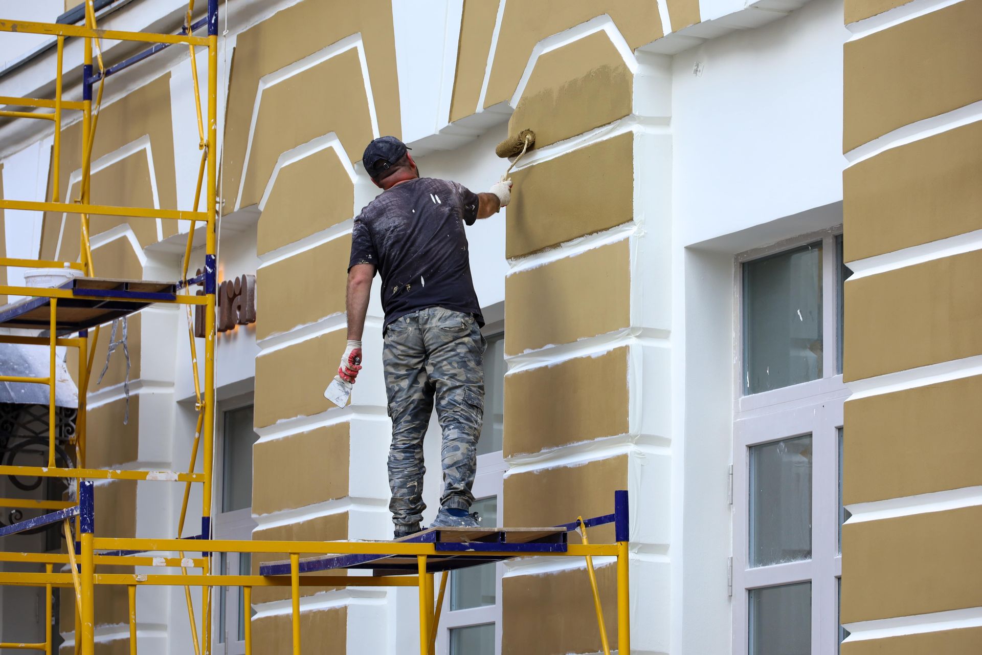 Man painting building facade from scaffolding, using roller, gold and white colors.