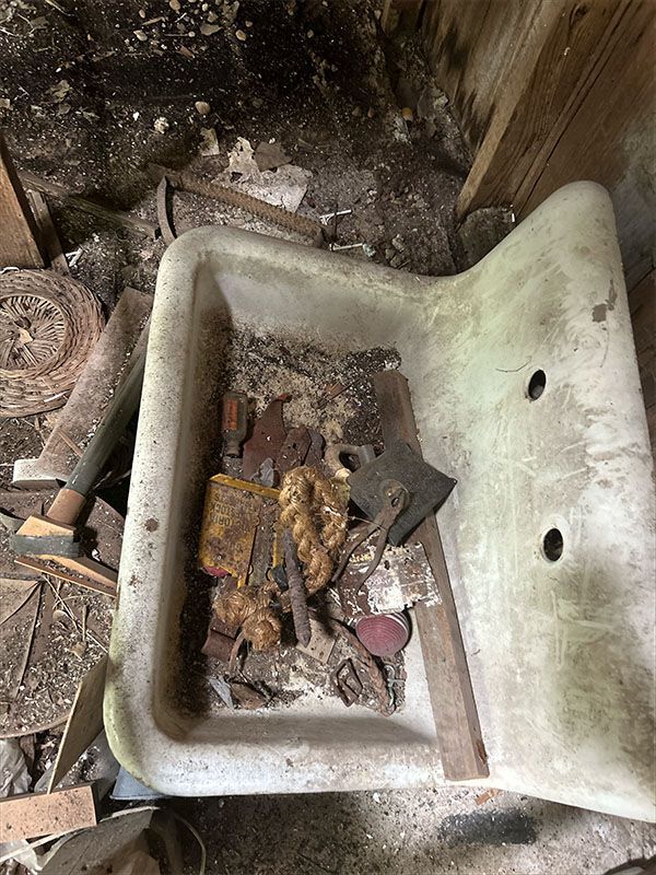 An old, stained white porcelain sink filled with debris including an ax, rope, and wood, in a decaying setting.