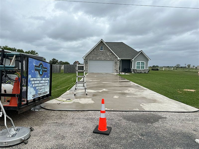 A concrete driveway being pressure washed in front of a house with a truck, ladder, and safety cone present.