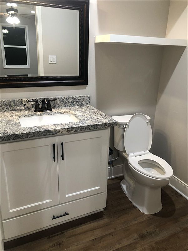 White bathroom with a toilet, vanity, and mirror. Granite countertop, black fixtures. Dark wood-look flooring.