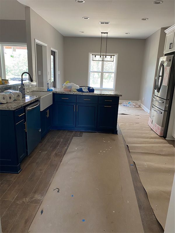 Kitchen with blue cabinets, island, and unfinished floors. Gray walls, a window, and a light fixture are also visible.
