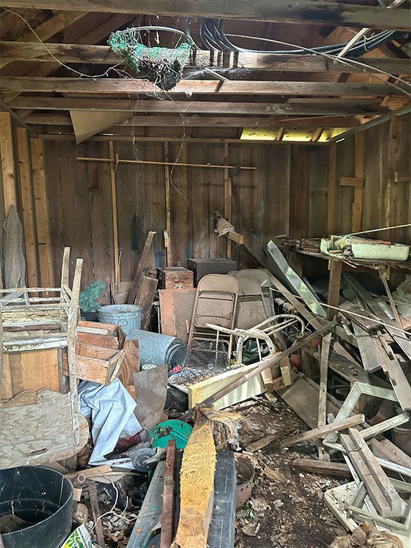 Cluttered wooden shed interior with old furniture, tools, and debris.