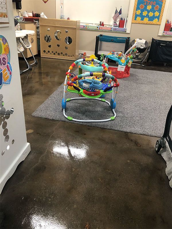 A baby play gym with toys sits on a gray rug inside a brightly lit daycare room.