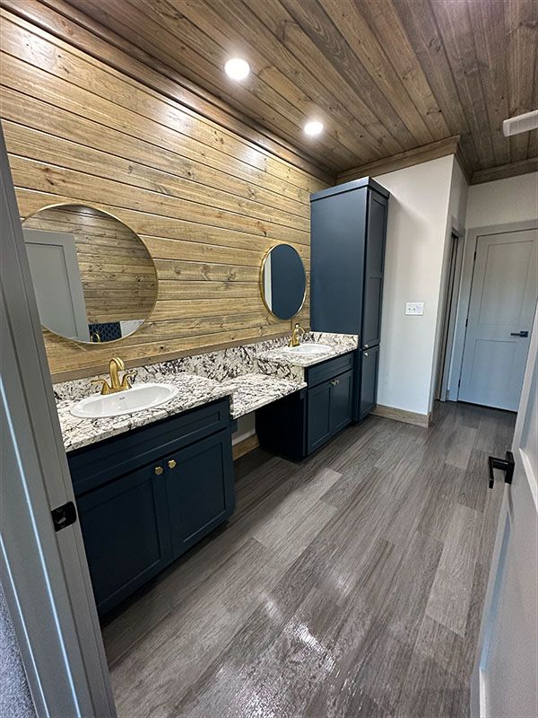 Bathroom with wood walls/ceiling, blue cabinets, marble countertops, and two round mirrors.