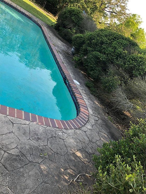 Poolside view with turquoise water, red brick edging, and stone patio surrounded by greenery.