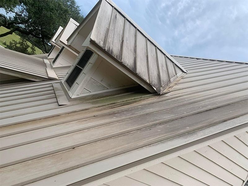 Light-colored metal roof with dark streaks and three small, triangular dormers under a cloudy sky.