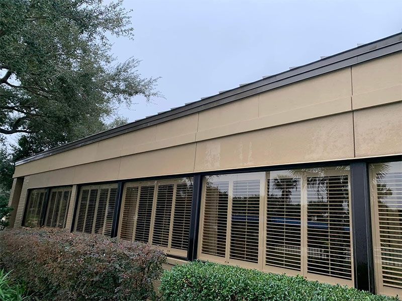Tan building with shuttered windows and dark trim, obscured by rain, trees in the background.