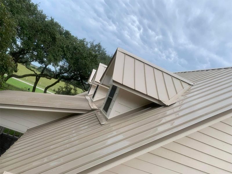 Tan metal roof with three vertical, angled dormers against a cloudy sky, and green trees in the background.