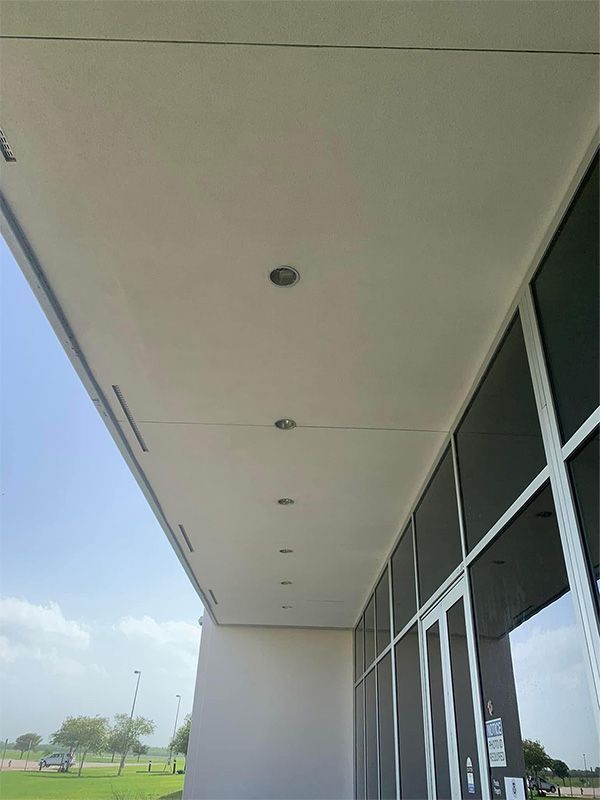 Exterior view of building with recessed lights in the soffit, next to large windows and a blue sky.