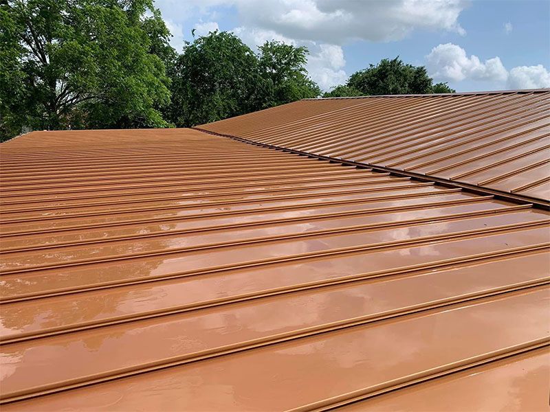 Copper-colored metal roof with rain drops, reflecting the sky and trees on a partly cloudy day.
