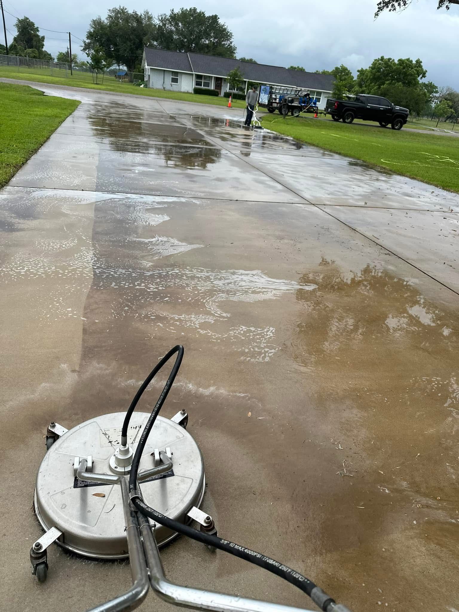 A person pressure washing a wet concrete driveway in front of houses on a cloudy day.