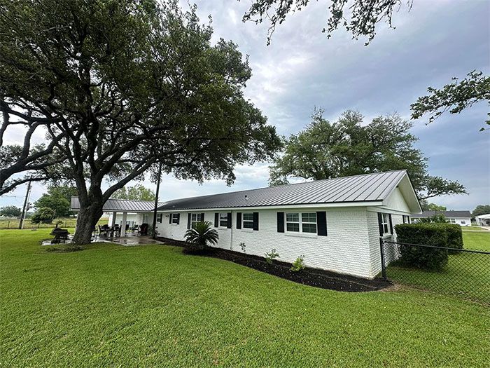 White brick ranch-style home with a metal roof, surrounded by a green lawn and a large tree. Cloudy sky.
