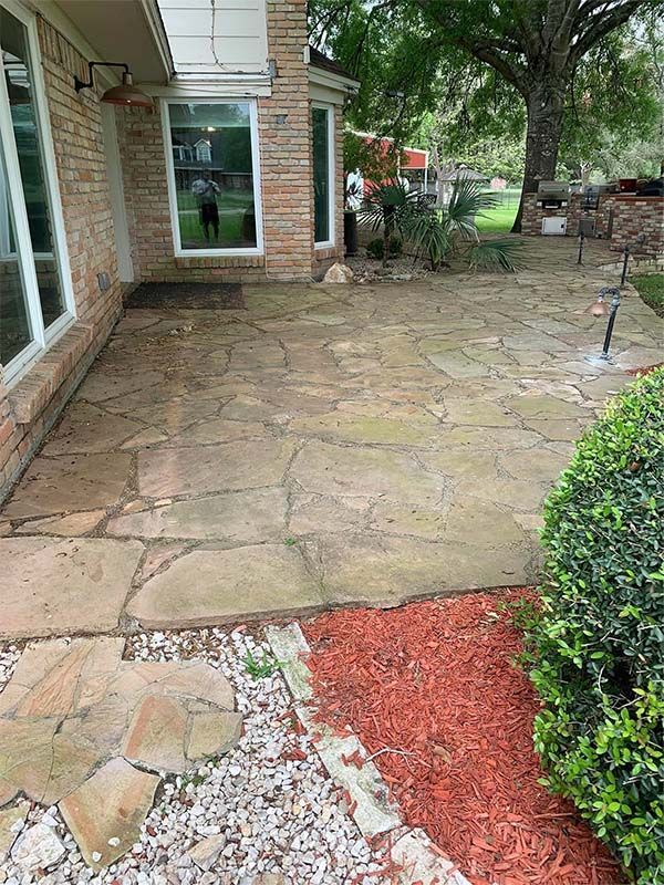 Stone patio beside a brick house, with landscaping and a large tree in the background.