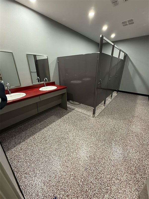 Public restroom with sinks and stalls. Grey walls, red countertop, speckled floor.