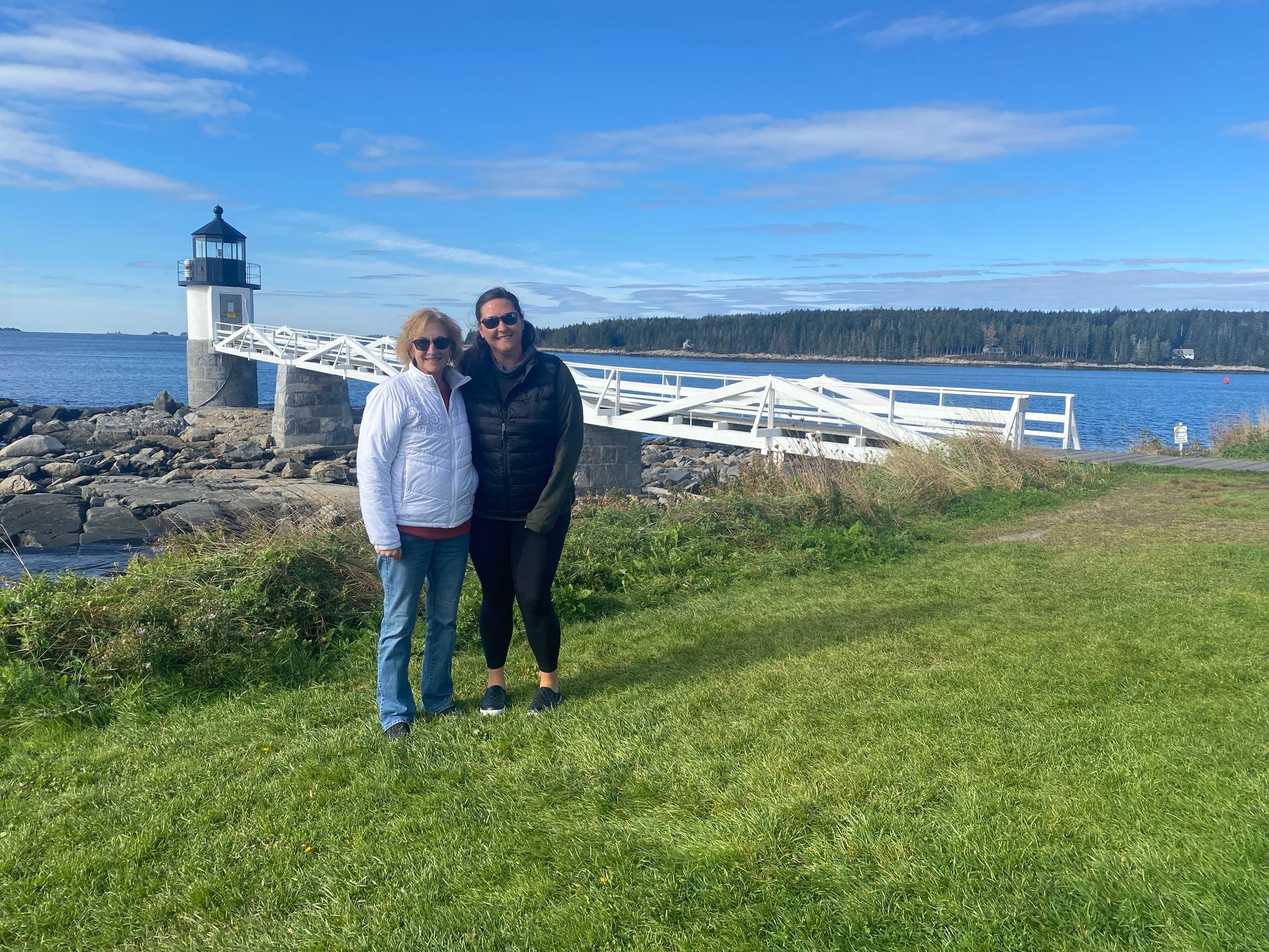 Two women stand in front of a lighthouse on a sunny day.