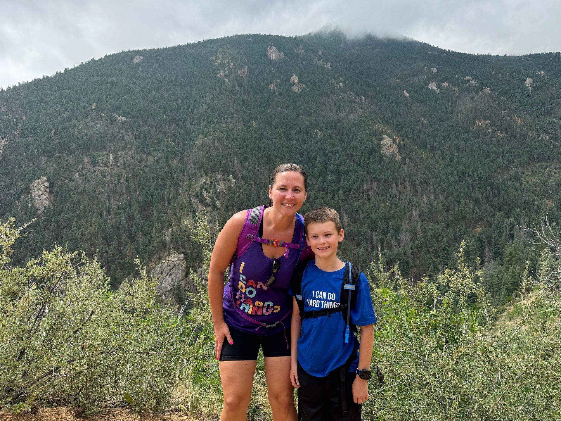 Woman and boy pose in front of a mountain covered in pine trees on a cloudy day.