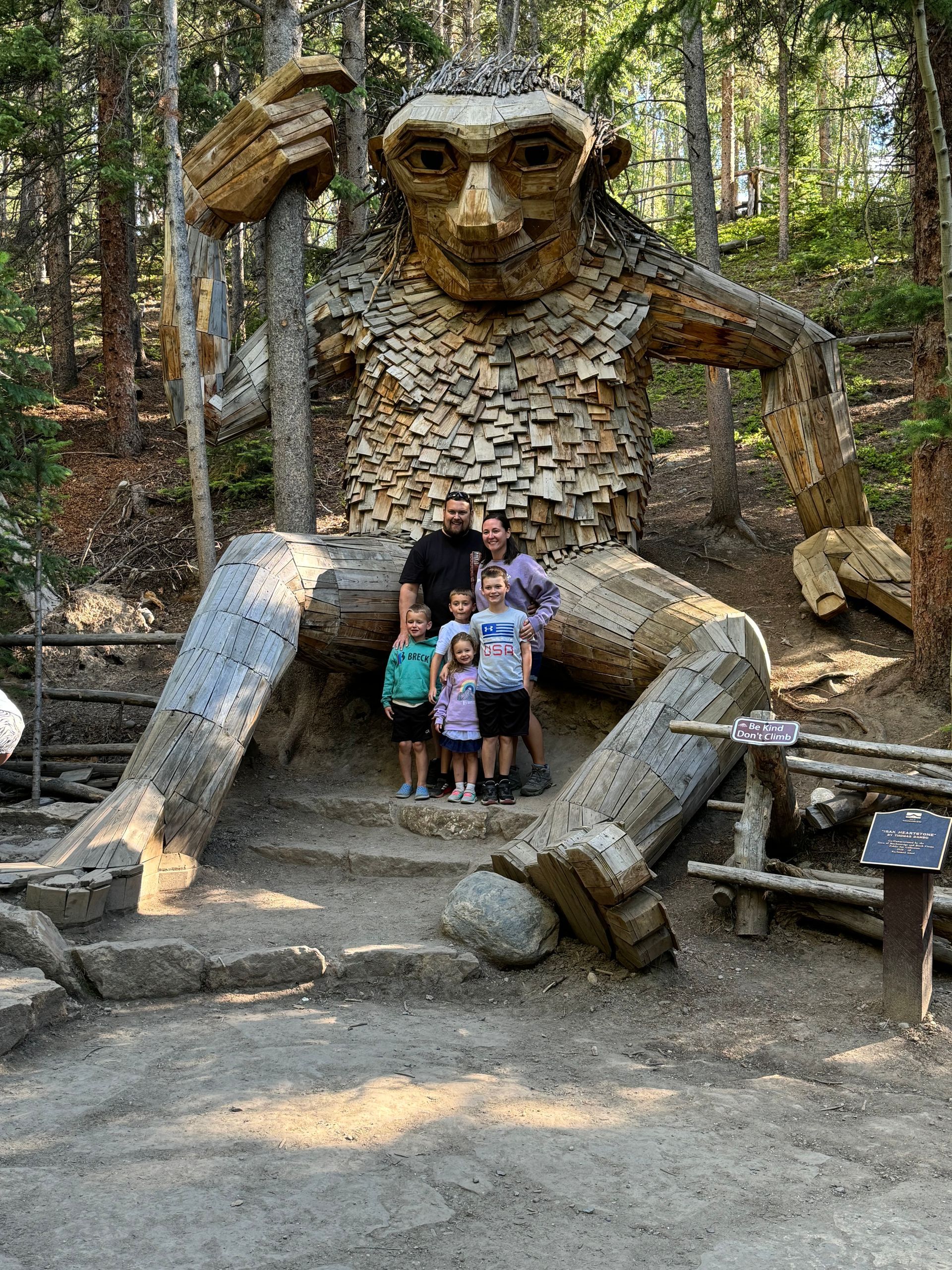 Family poses in front of a giant wooden troll sculpture in a wooded area.