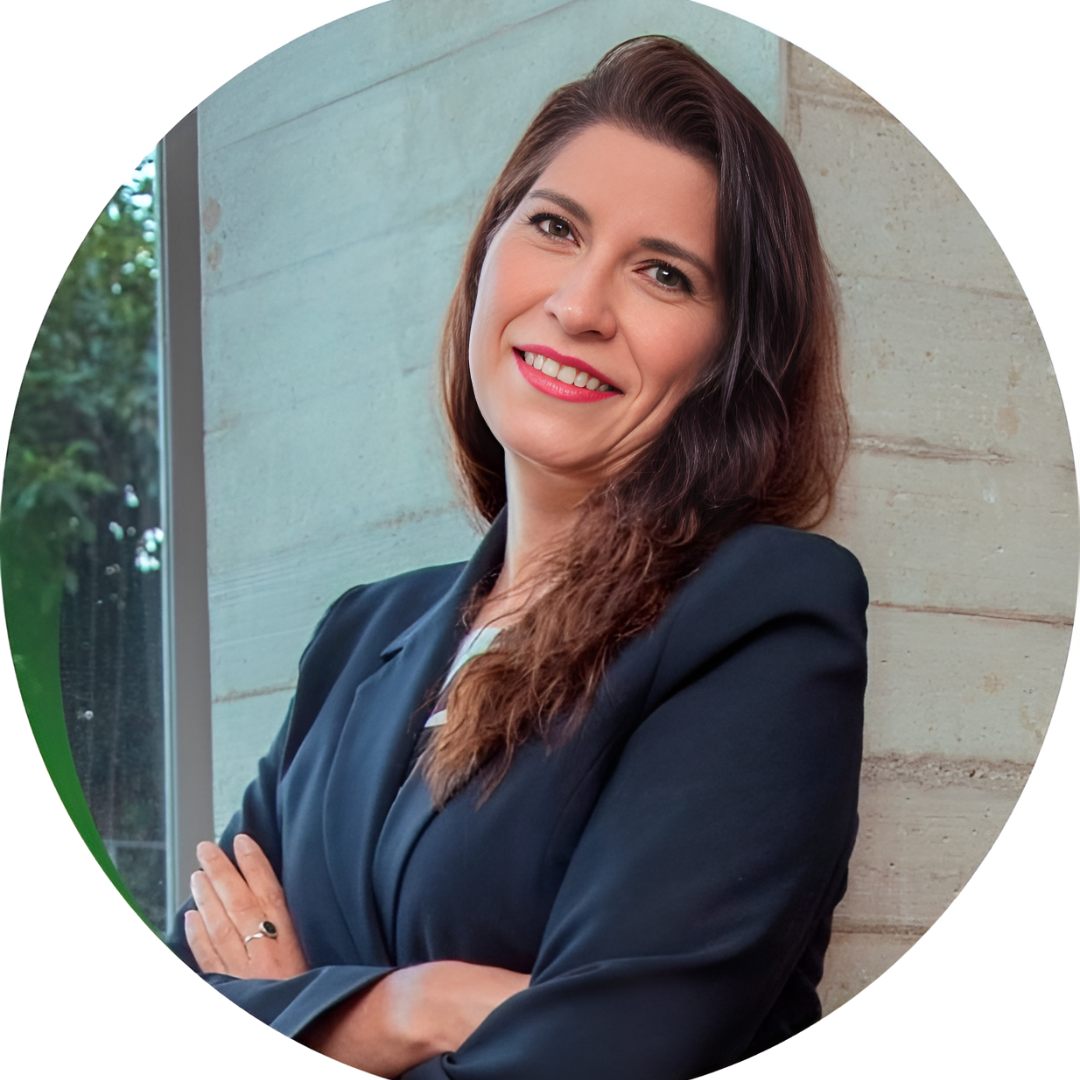 Una mujer con blazer azul marino sonríe con los brazos cruzados, apoyada contra una pared de hormigón cerca de una ventana.
