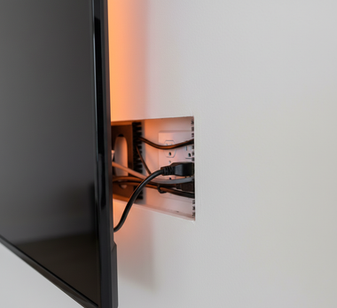 A TV is mounted on a wall next to a recessed cable management box with power cords and a surge protector.