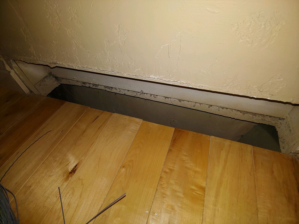 Open floor vent cut into a wooden floor beside a beige wall