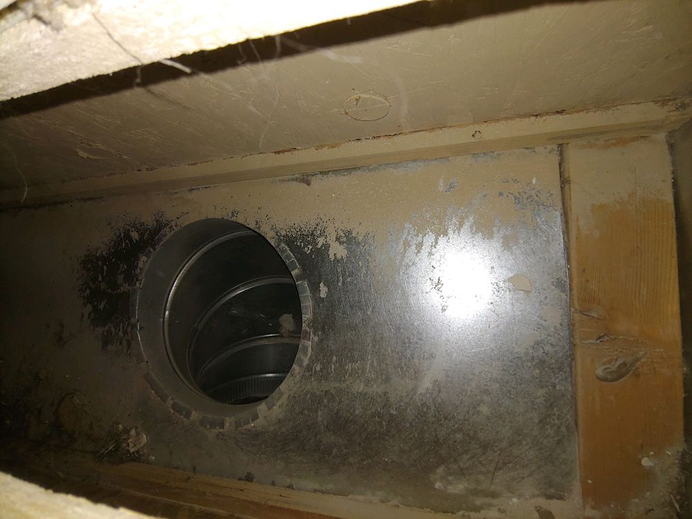 Dusty metal vent duct inside a wall opening with a round pipe visible.