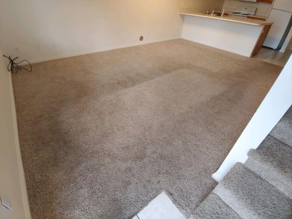 Empty beige-carpeted living room with stairs and a small kitchen counter area.