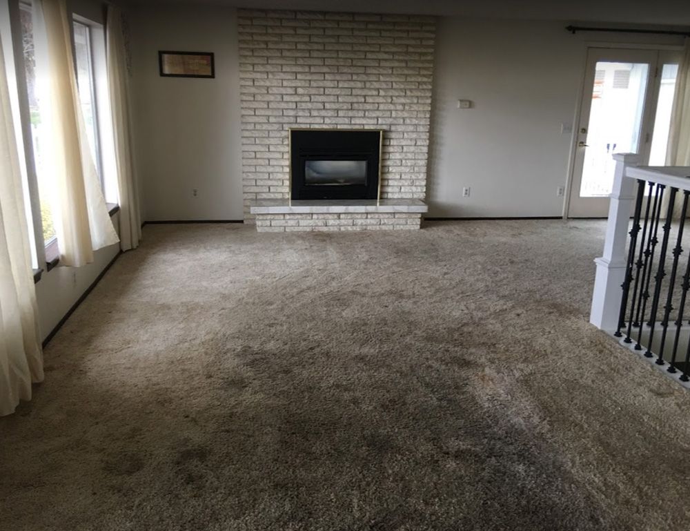 Empty carpeted living room with a white brick fireplace and large windows.