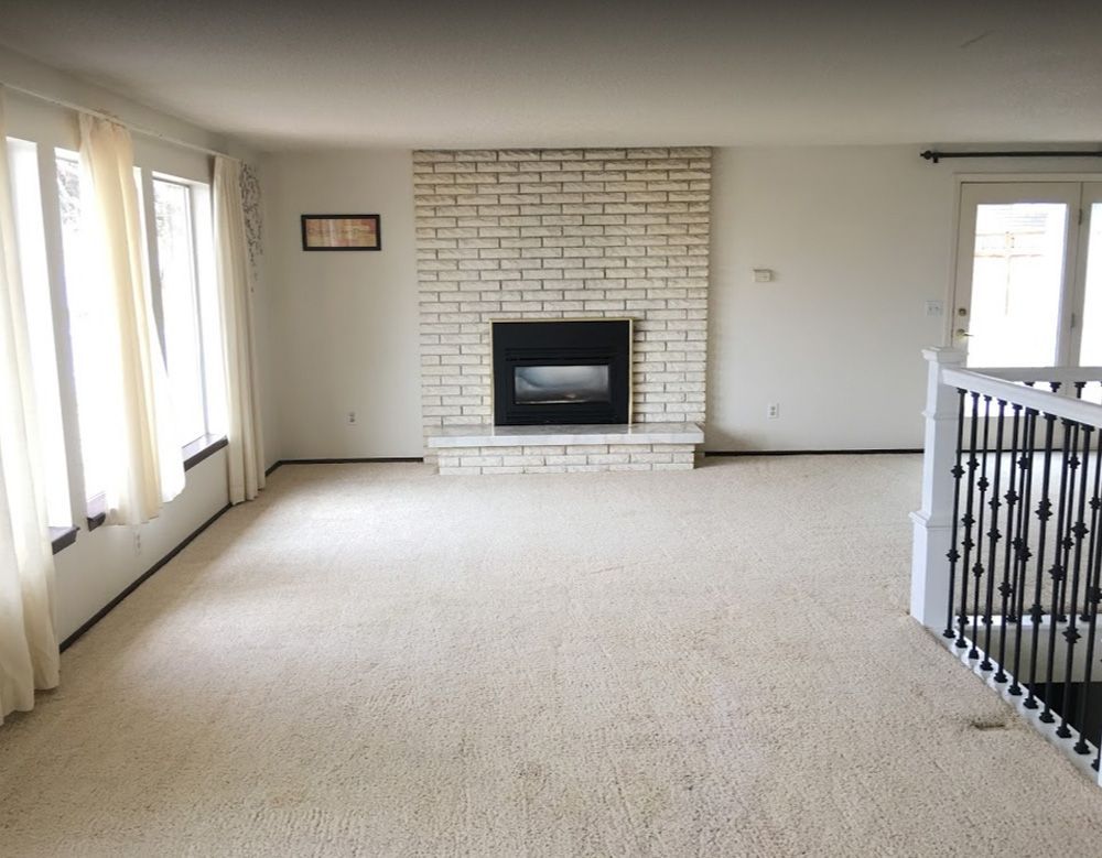 Empty carpeted living room with white brick fireplace and large windows along one wall
