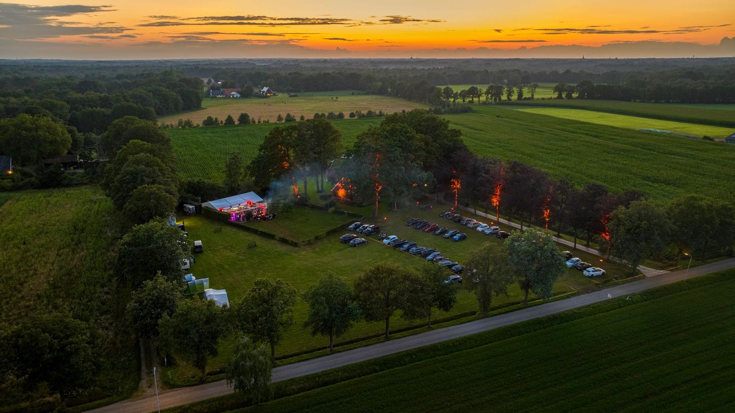 Luchtfoto van een buitenevenement in de schemering; bomen met verlichting, een veld en een gebouw onder een oranje hemel.