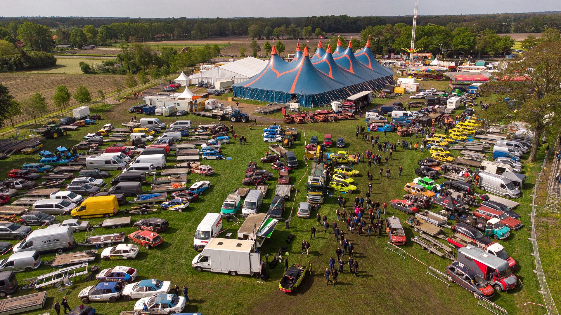 Luchtfoto van een festival met auto's, busjes en een grote circustent op een veld.