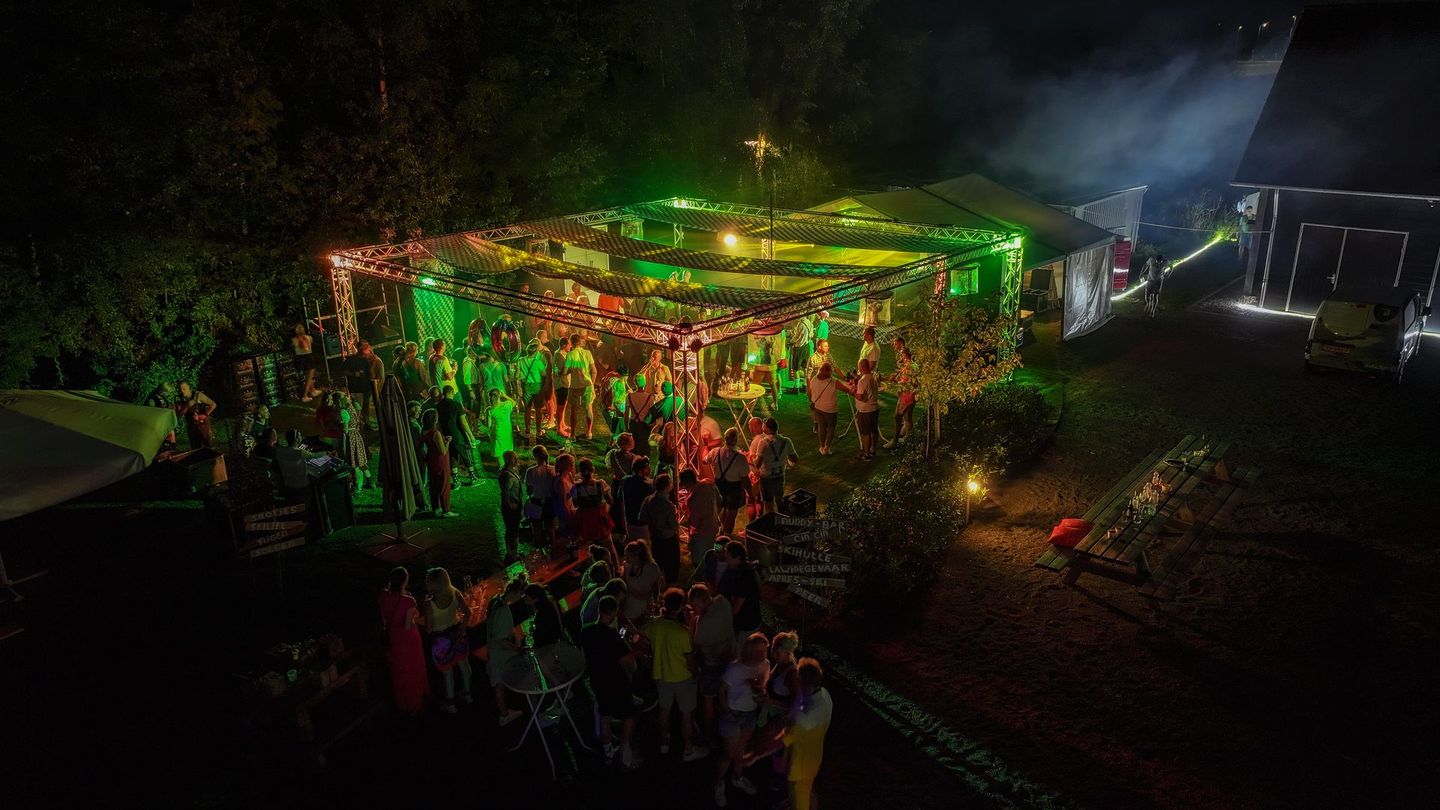 Een avondlijk buitenfeest met een menigte onder een verlichte luifel; groene, rode en witte lichten.