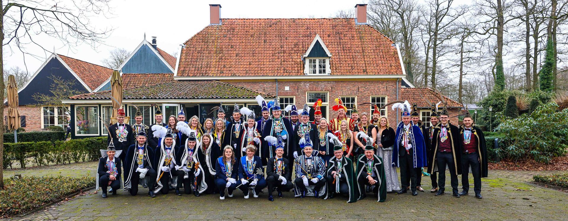 Een groep mensen in kostuums poseert voor een huis met een rood dak.