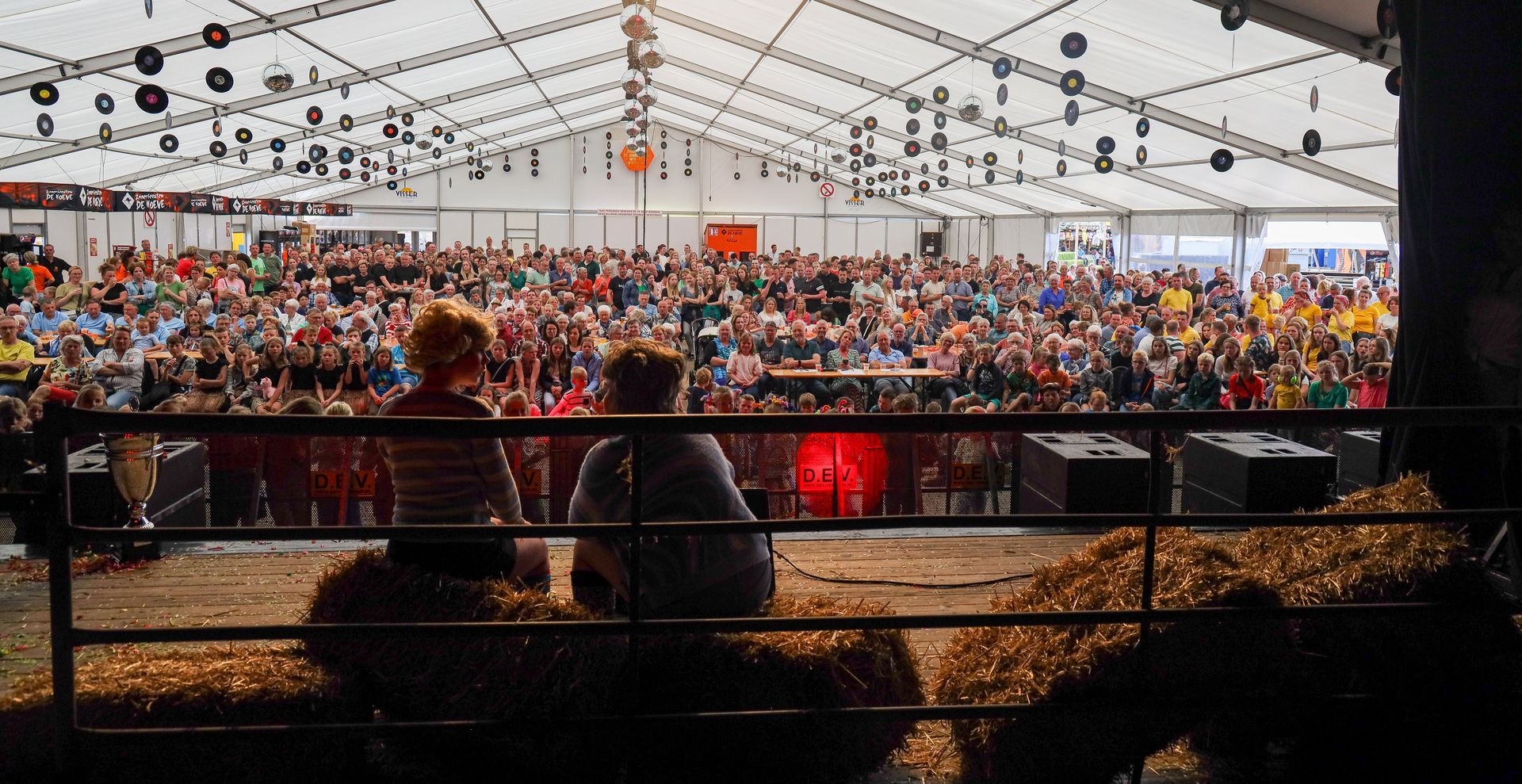 Twee mensen op hooibalen kijken naar een grote menigte in een tent op een festival.
