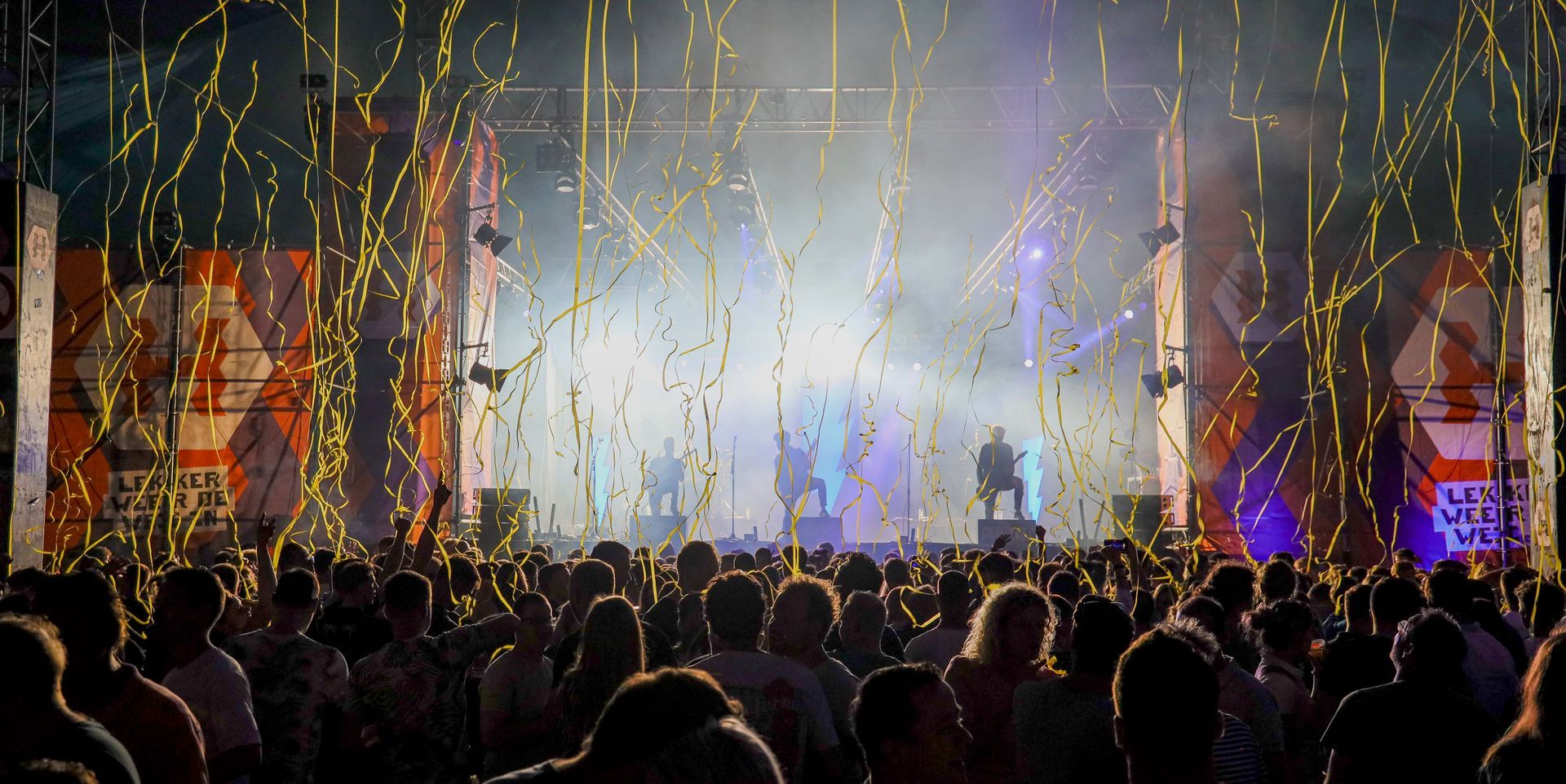 Concertpodium met artiesten, publiek, vallende gouden confetti en felle lichten.