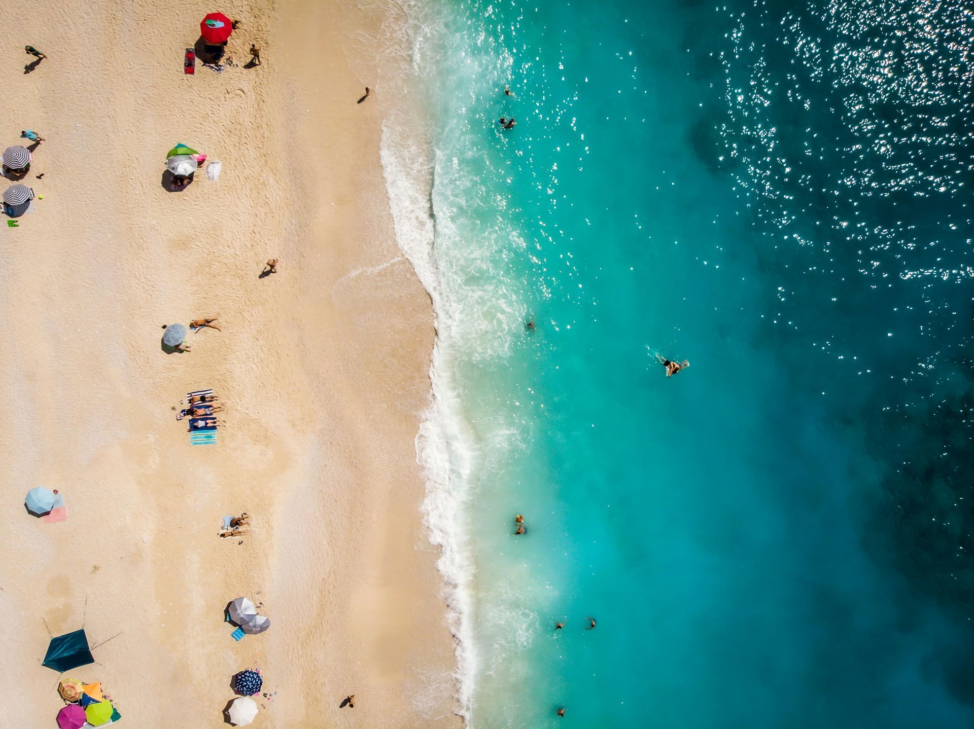 Een strand vol mensen, parasols op het zand, turquoise water met zwemmers.