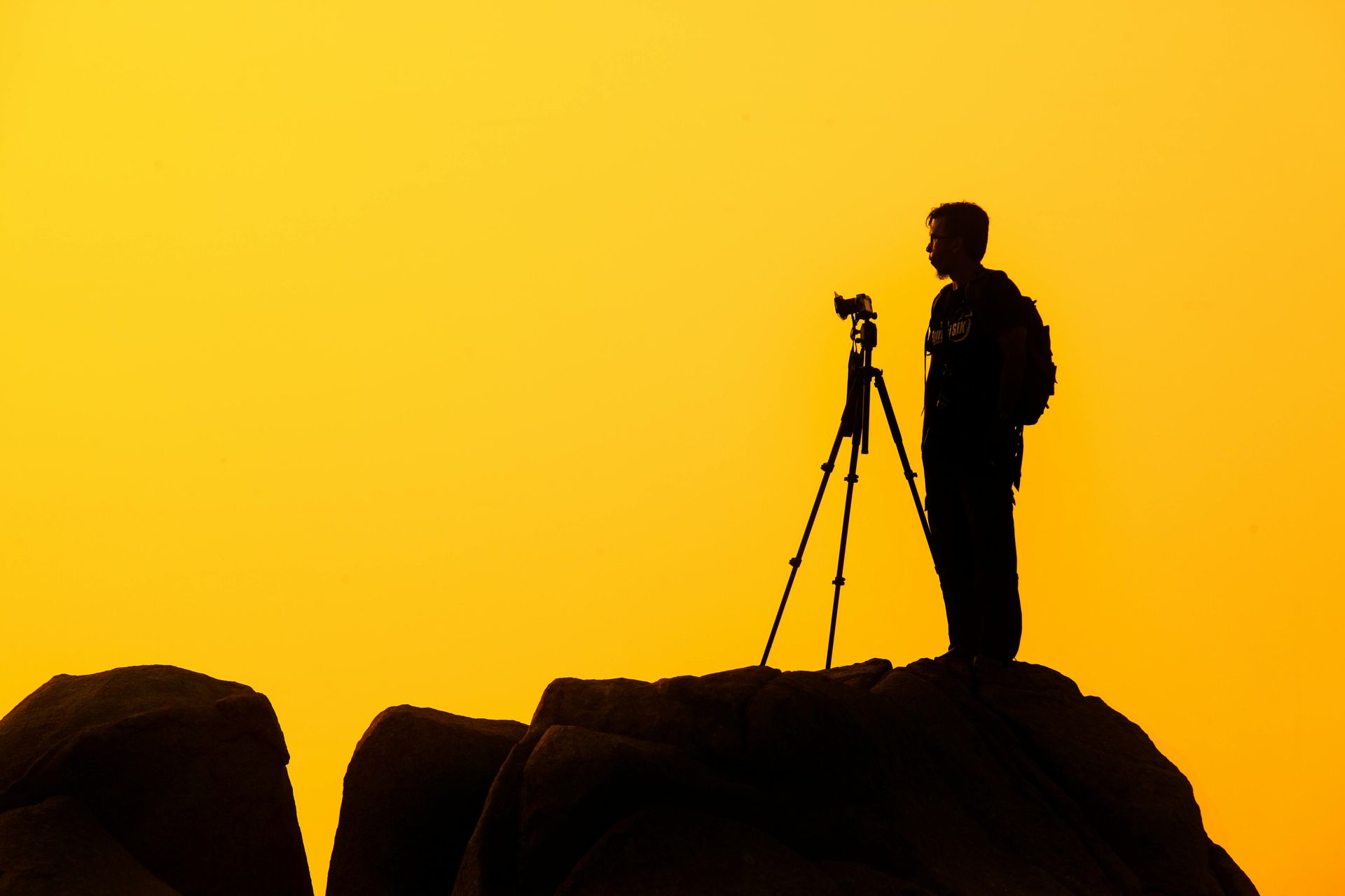 Silhouet van een fotograaf op een rots met statief, tegen een heldergele hemel.