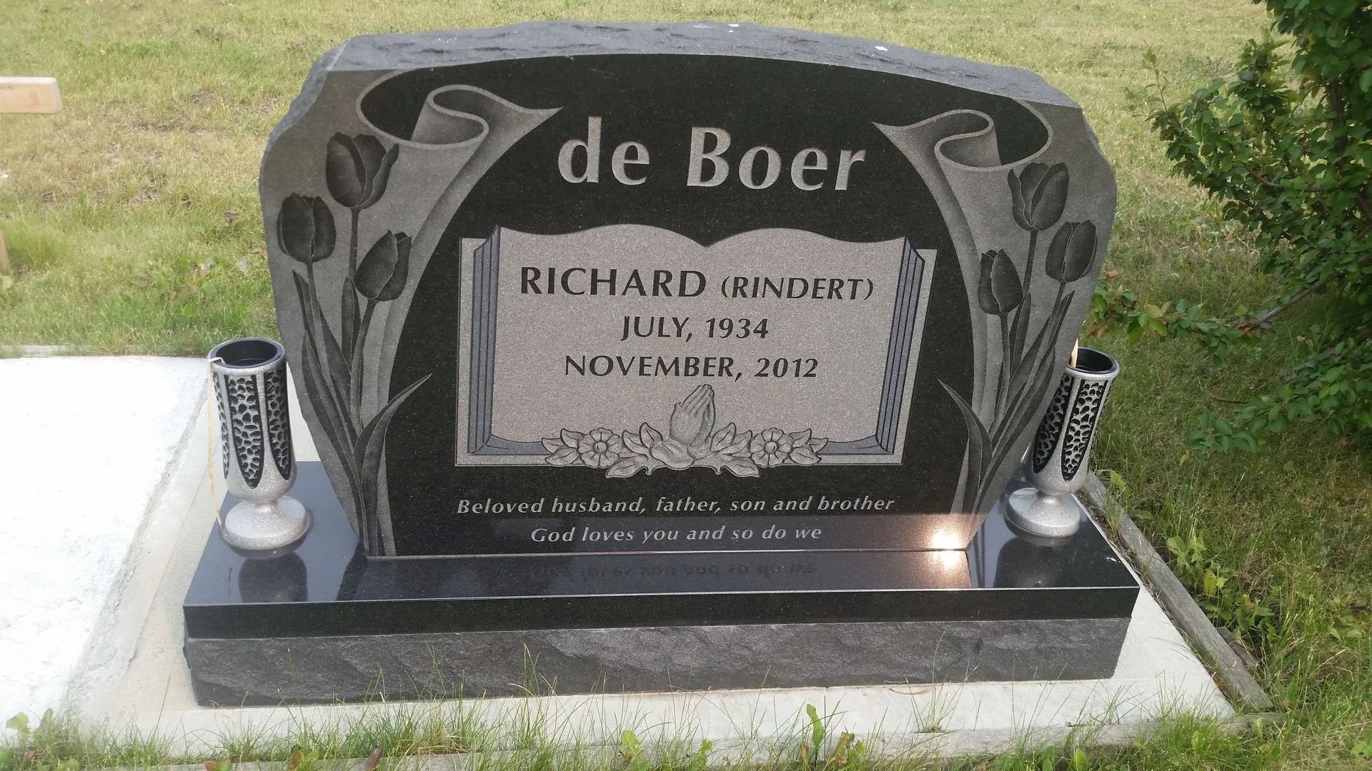 Black granite gravestone with etched design and flowers, reading “de Boer” in a grassy cemetery.