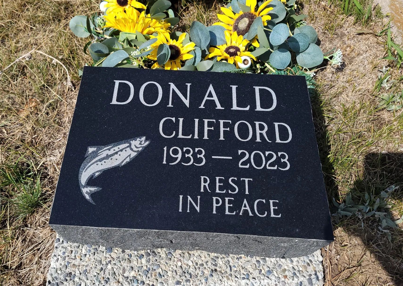 Black headstone with name Donald Clifford, years 1933-2023, and inscription 'Rest in Peace', decorated with flowers