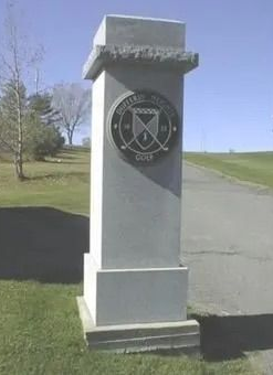 Stone memorial pillar with a circular emblem beside a paved road and grassy lawn