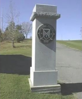 Stone memorial pillar with a circular emblem beside a paved road on a grassy hillside