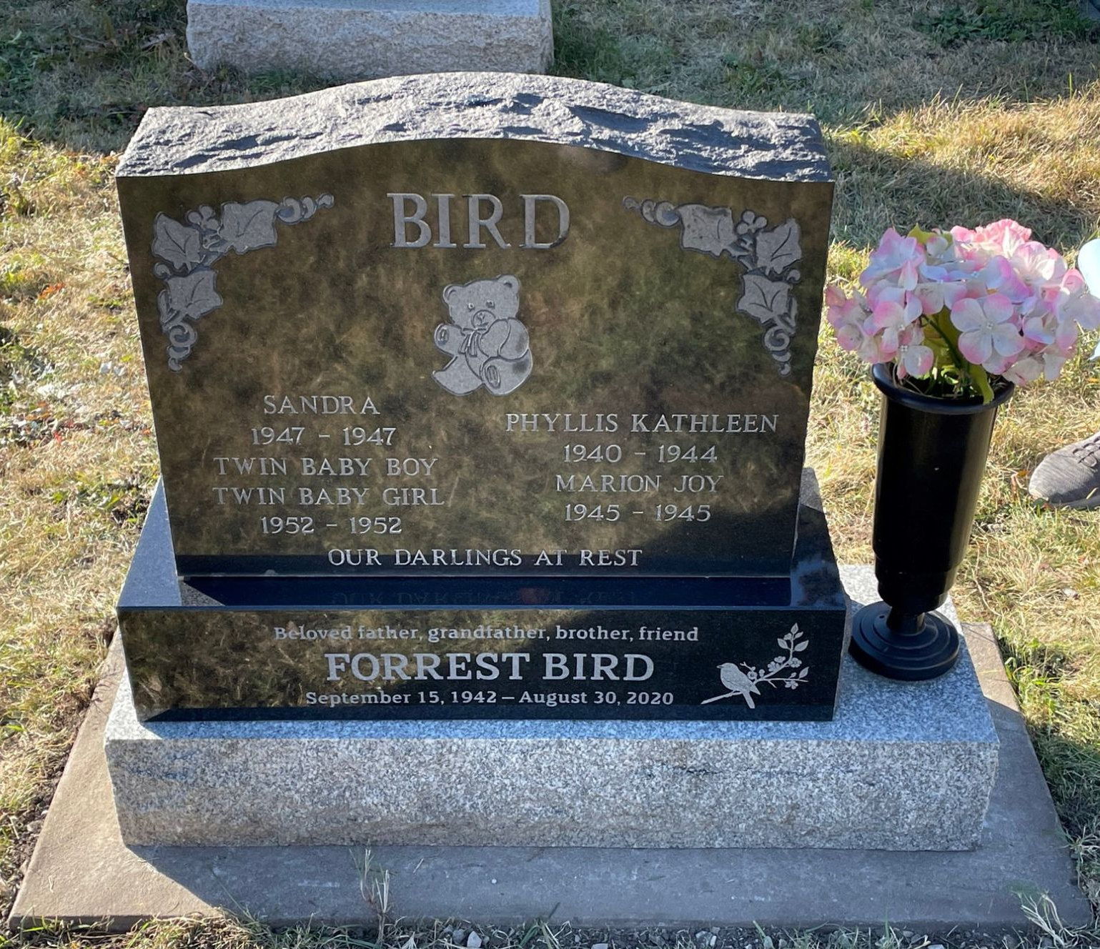 Gravestone for Forrest Bird with flowers in a black vase beside it in a cemetery.
