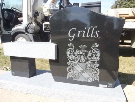 Black headstone with engraved name 'Grills' and heraldic crest, displayed next to white bench in cemetery