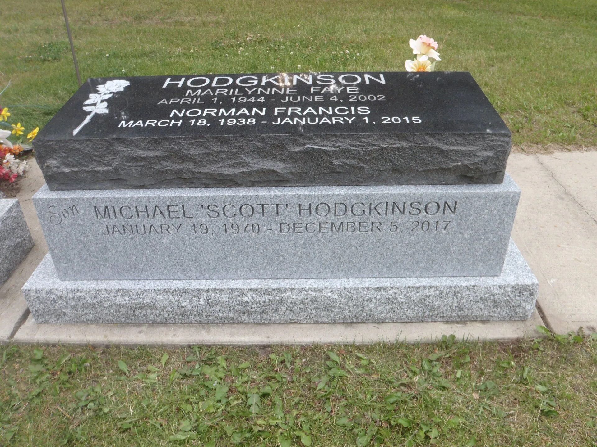 Black granite memorial headstone with engraved names, dates, and flowers on a grassy lawn.