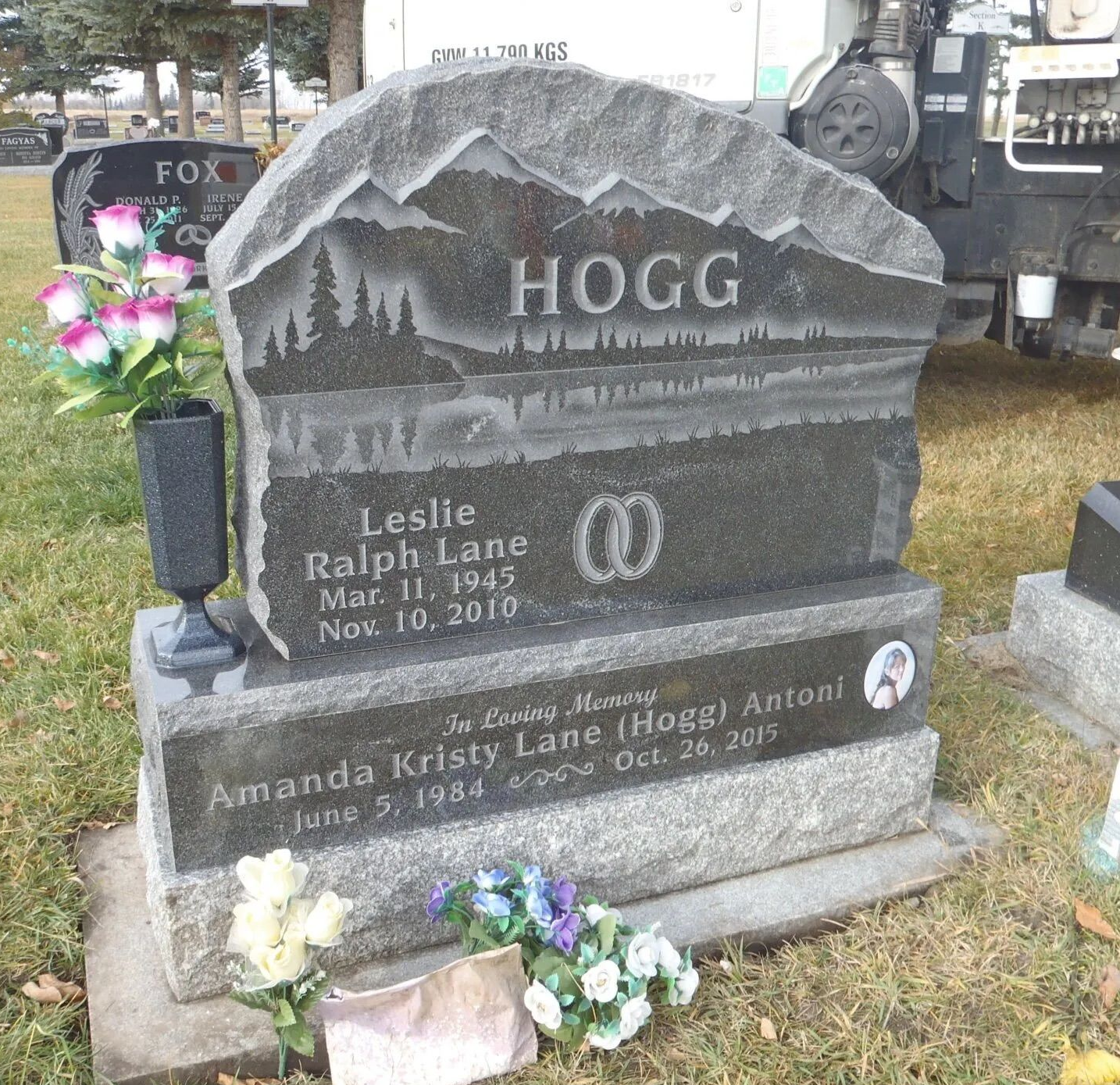 Gravestone with “Hogg” engraved, mountain scene, and flowers at the base
