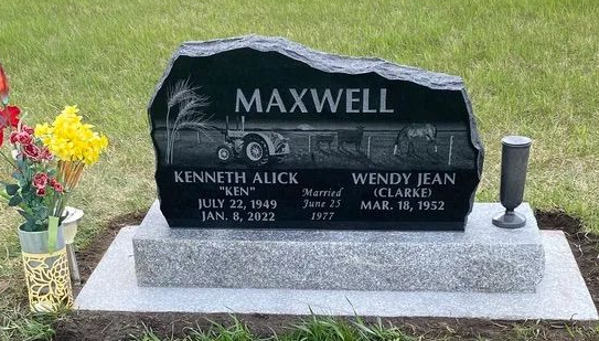 Black Maxwell gravestone with engraved names, set on a gray base in a grassy cemetery with flowers on the left