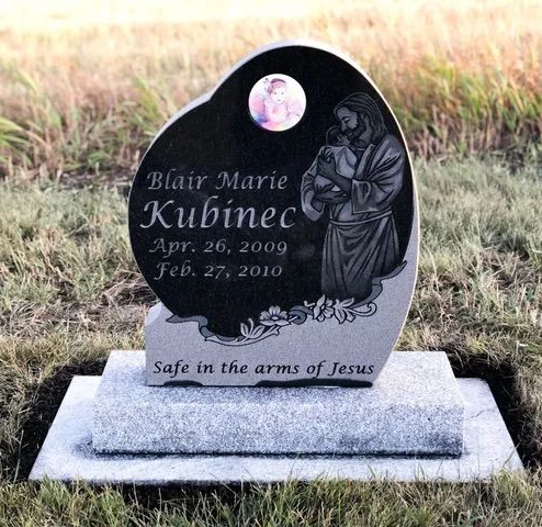 Black gravestone with angel engraving and inscription, “Safe in the arms of Jesus,” in a grassy cemetery