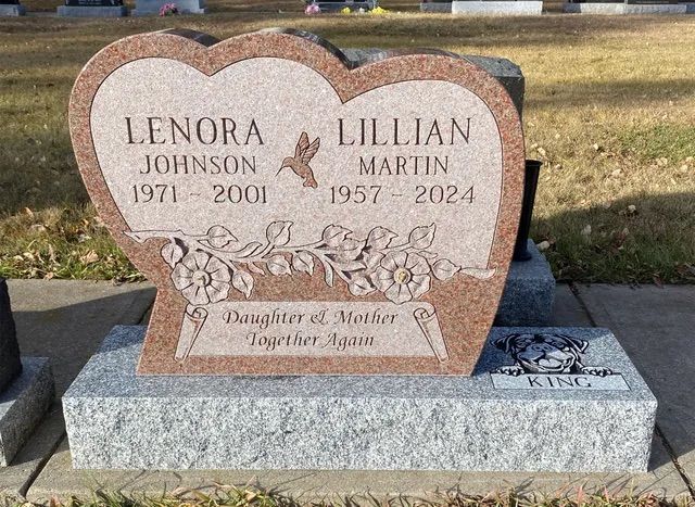 Granite memorial headstone with pink heart-shaped plaques and flowers engraved, in a cemetery.