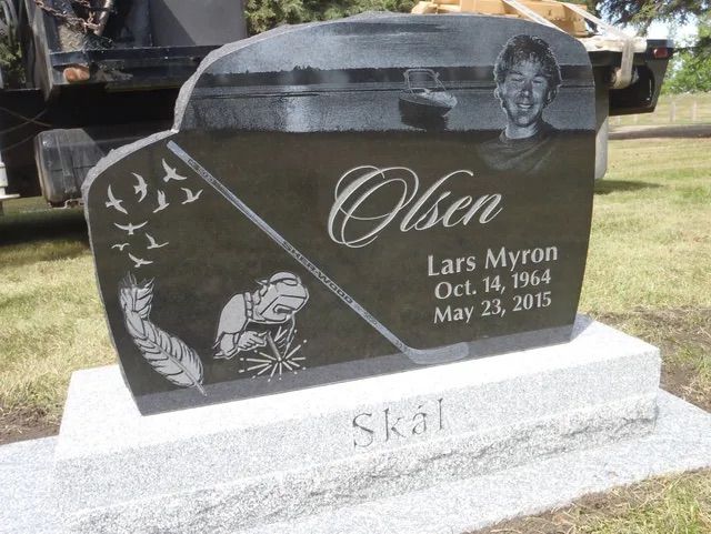Black granite memorial gravestone with engraved birds, flowers, and name “Olsen” in a cemetery.